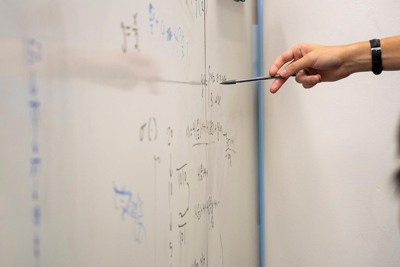 A hand on the left of the photos points at calculations written on a dry erase board.