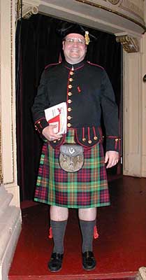 A man wears full Scottish regalia, including a green and red plaid kilt, black jacket, jaunty hat, knee socks and garter flash.