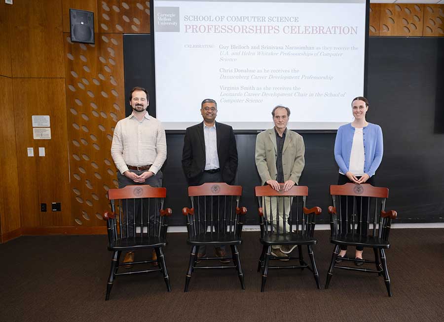  Three men and a woman pose behind physical chairs in front of a screen welcoming attendees to the professorship celebration.