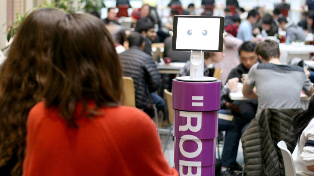 A robot with an iPad head and purple cylindrical body interacts with a woman in a crowded campus dining area.