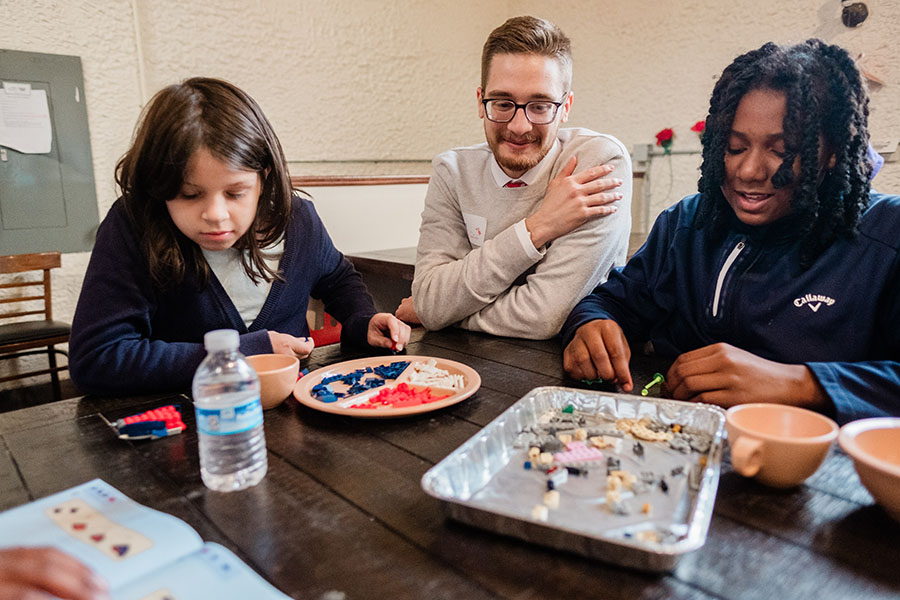 Two young women sit at a table building LEGO kits while a male instructor looks on.