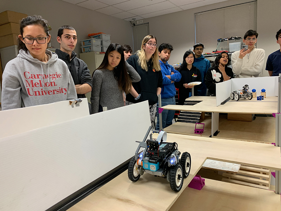 A group of 10 students gather around posterboard model of a building that is being navigated by a small car-like robot.