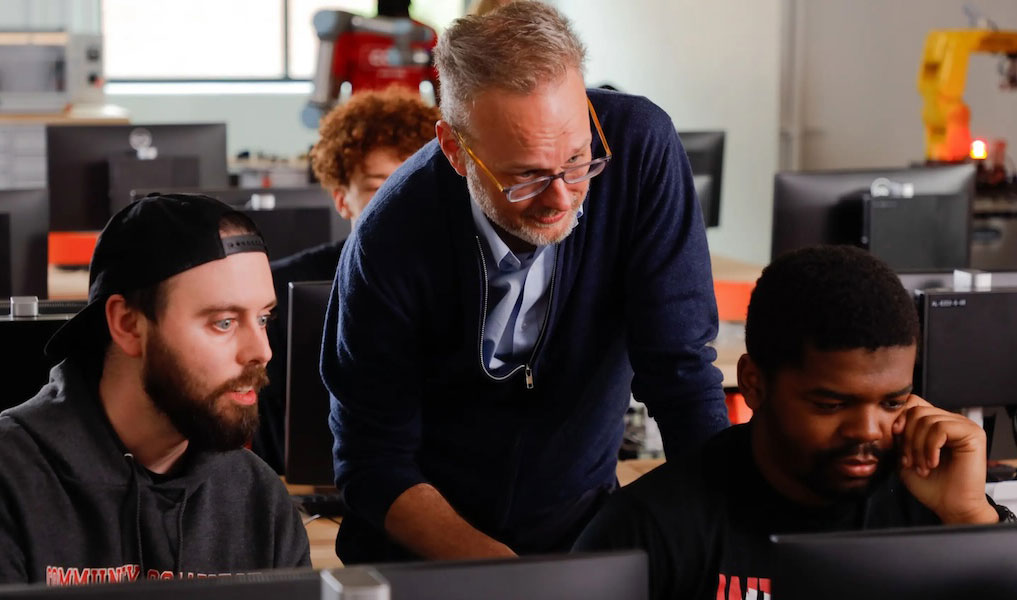  Two students work at laptops while an instructor looks over their shoulders.