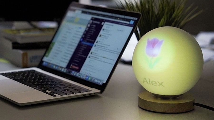 A yellow lamp on the right displays a flower emoji and the name Alex while a laptop is shown out of focus in the background.