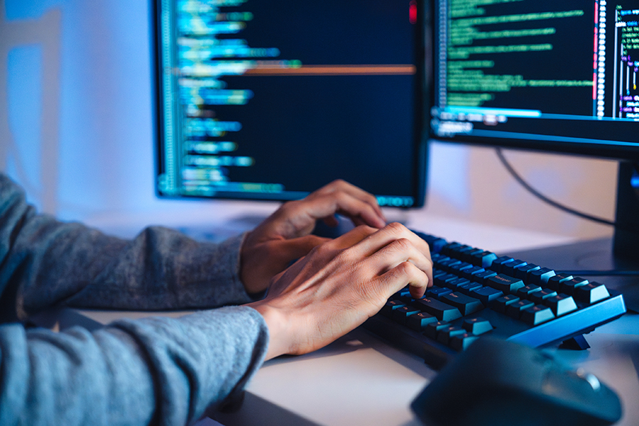  Humans hands type on a keyboard while multiple monitors appear blurred in the background.