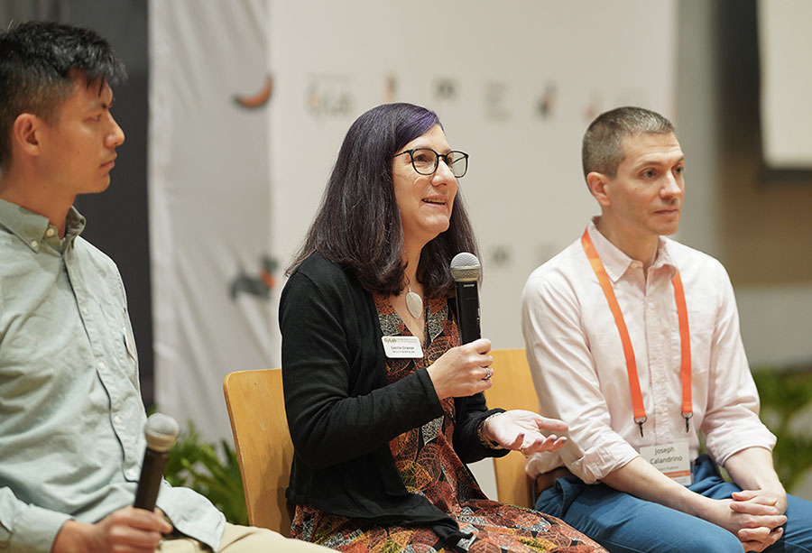  A woman sitting between two men on a stage speaks into a microphone.