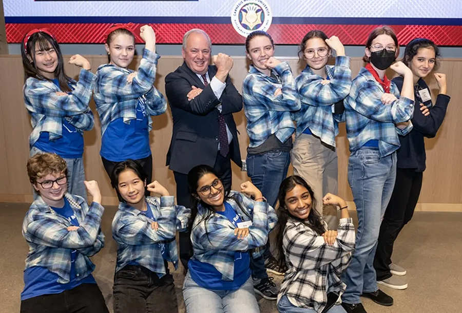 A middle-aged man in a business suit surrounded by young women dressed in Rosie the Riveter style plaid shirts and headbands gives the camera the Rosie the Riveter salute.