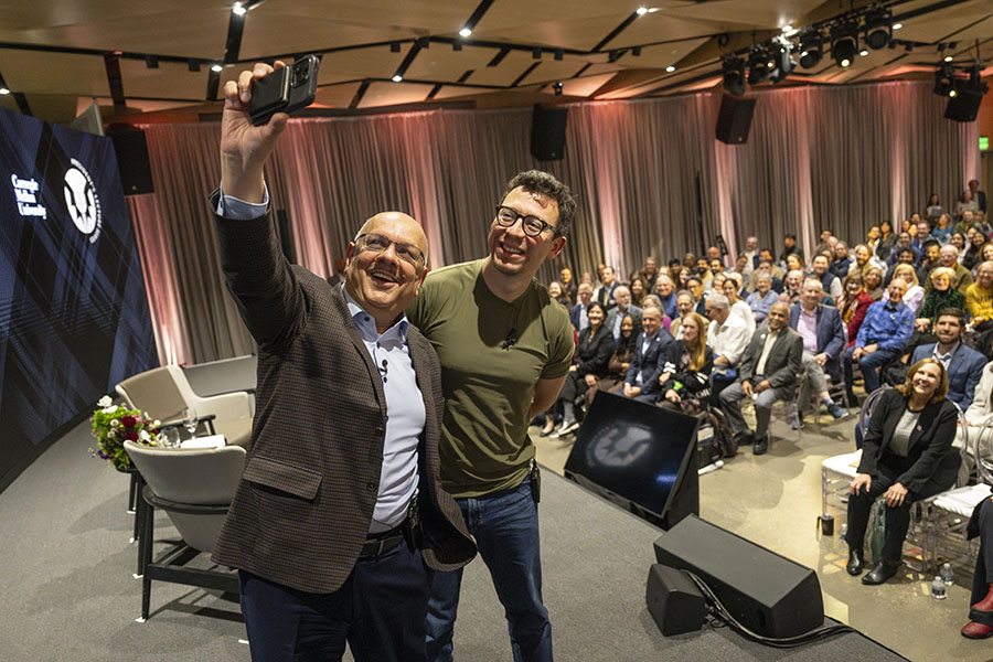  Two men on a stage take a selfie with the crowd behind them.