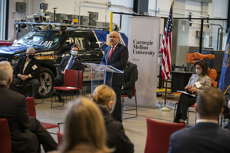 A man in a suit stands at a lectern in front of a Carnegie Mellon University sign. An audience is seated in the foreground, two men sit on his right in front of an SUV, and a woman sits on his left.