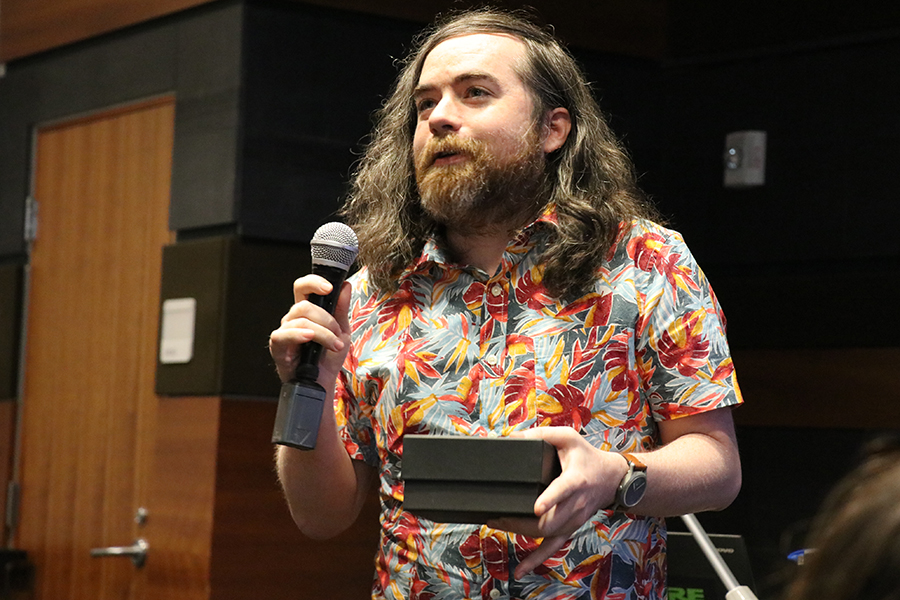 A man with long brown hair wearing a hawaiin shirt stands holds a microphone and accepts an award.