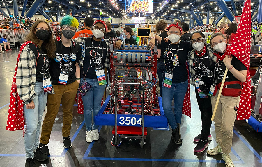 Six members of the Girls of Steel team, dressed in black tshirts with a Rosie the Riveter theme, pose with a robot that is shaped like a large shopping cart and is filled with red wires.