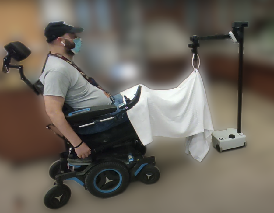 A man in a wheelchair uses a hands-free microphone and head-worn sensor to control a robotic hand that helps him adjust a blanket.