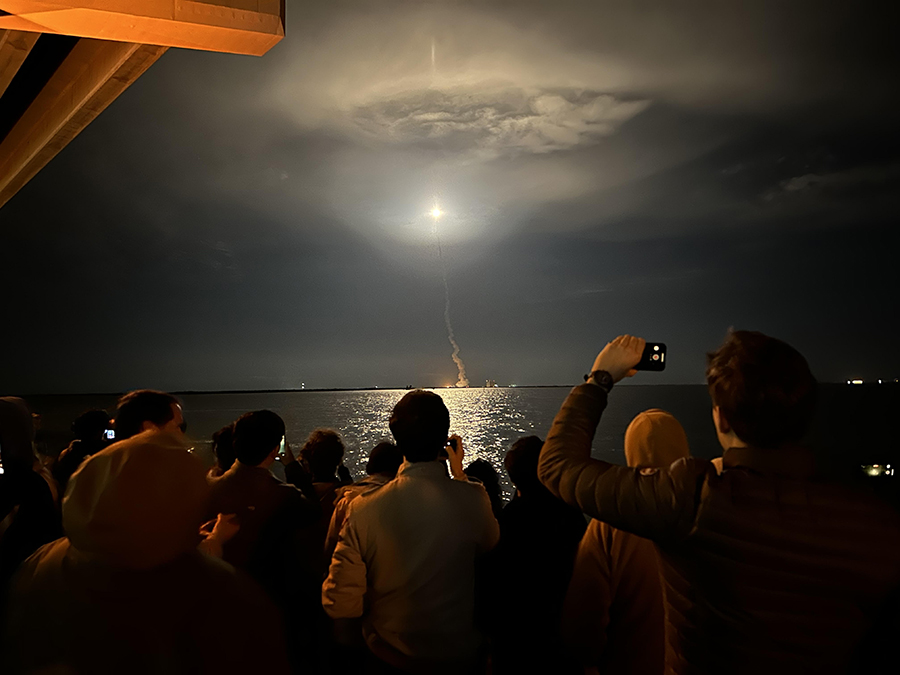 A rocket takes off in the background while a group of people, backs to the camera, watch and take photos.