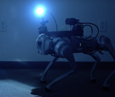  A four-legged robot with a camera mounted on its back walks in the dark with the help of a light strapped to its shoulders.