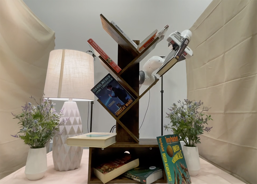  A robotic arm removes a book from a shelf that's surrounded by plants and a lamp.