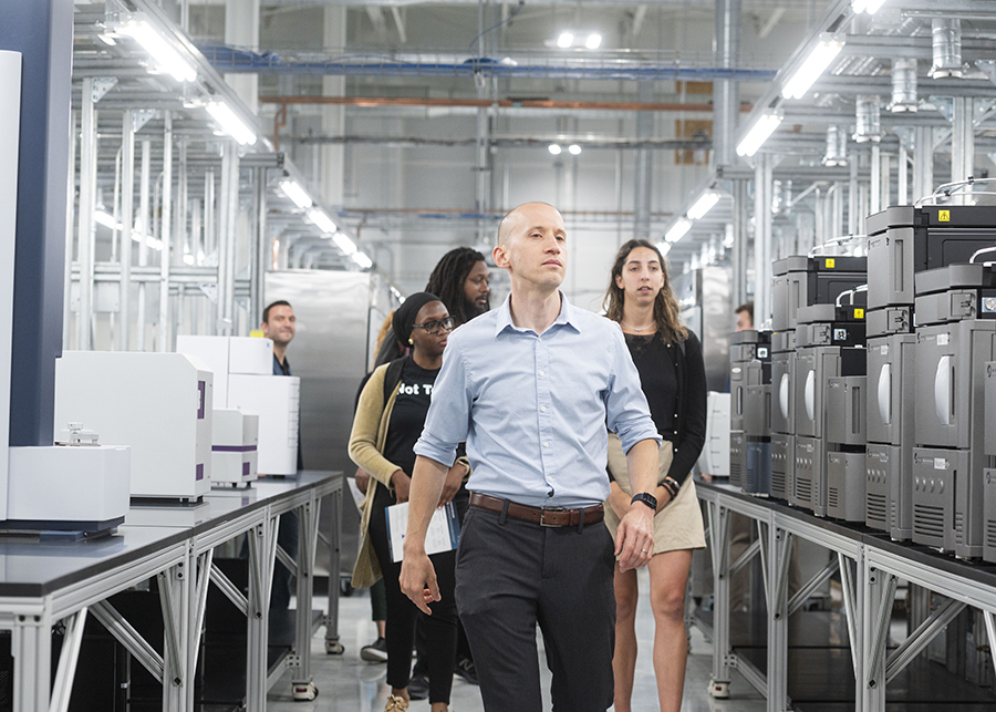 A man in light blue shirt leads a group of people walking down an aisle in a stark lab with servers and equipment on shelves, and industrial pipes in the background.