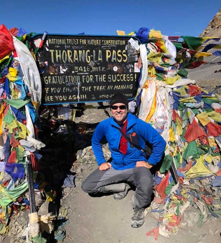 A man in a blue jacket and hiking gear kneels in front of the sign for Thorong La mountain pass.