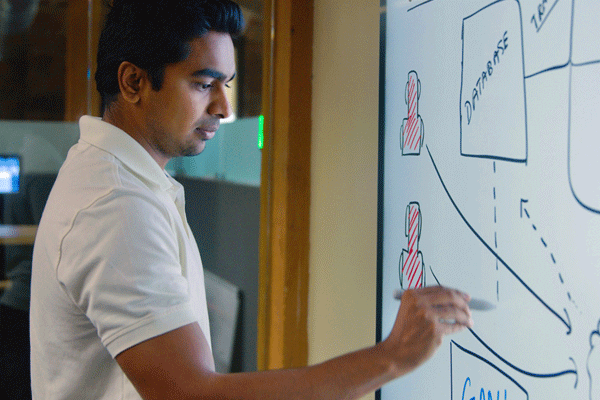 A man in a white polo shirt sketches on a dry erase board. 