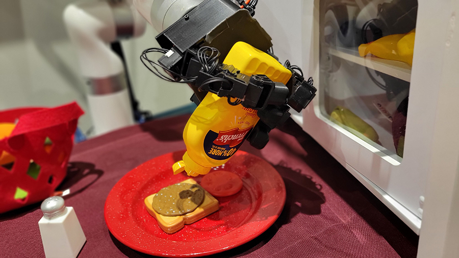 A robotic hand grips a yellow mustard bottle and tilts it over a toy hamurger patty on a slice of fake bread, which is sitting on a red plate.