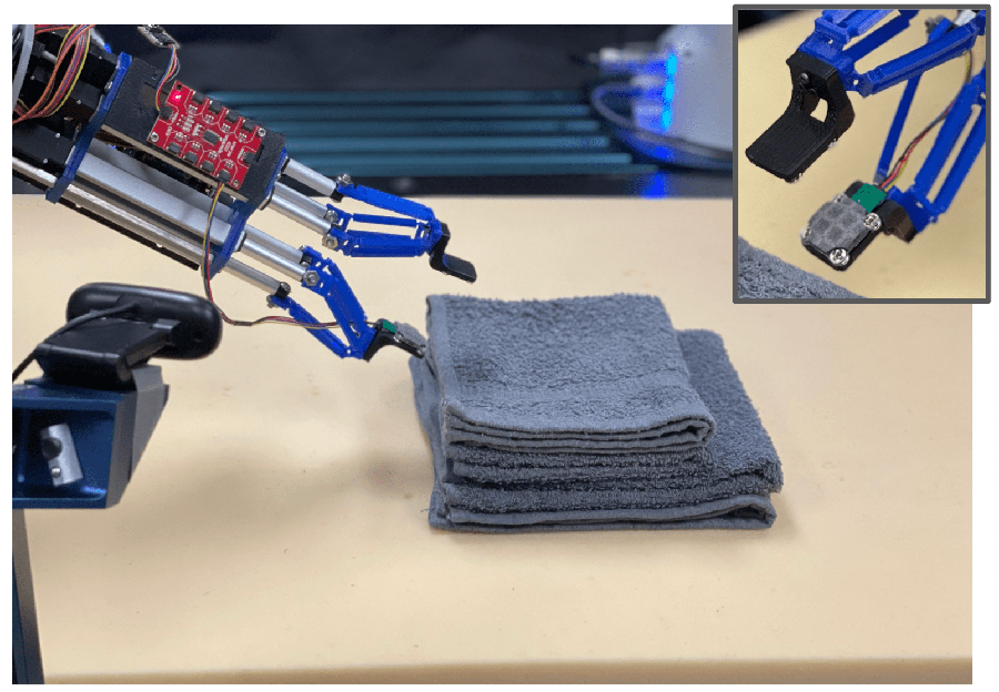 A robotic arm with two flat prongs representing the hand grasps a blue hand towel. An inset in the upper right corner shows a close up of the pronged fingers and the sensor strip attached to them.