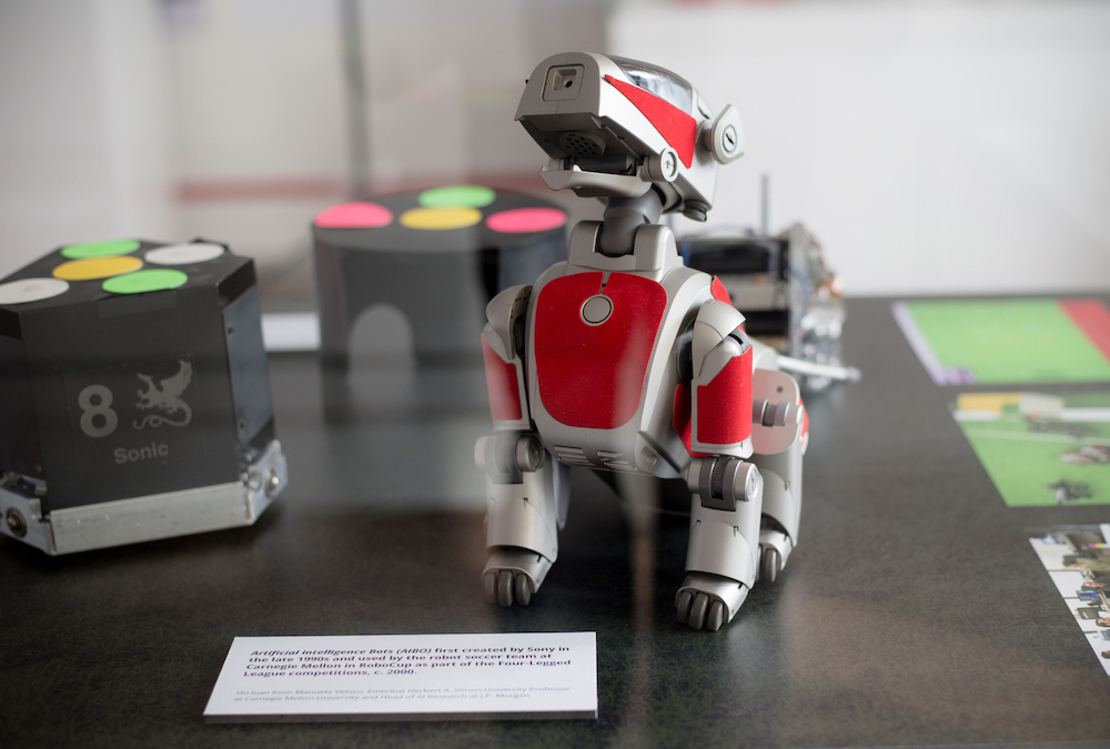 A silver and red robot dog sits on a black table, with two black cube robots in the background.
