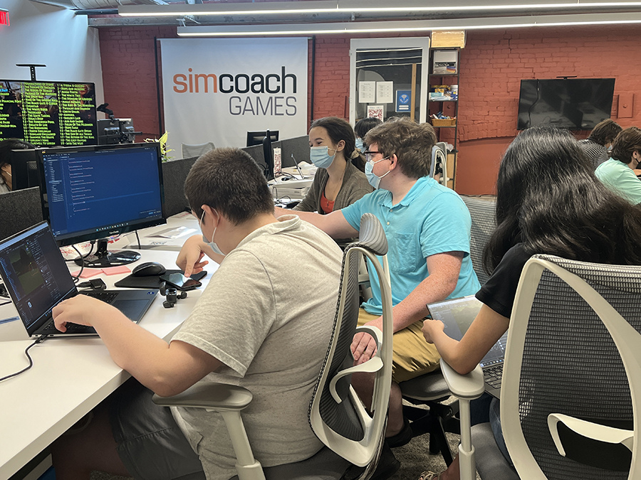 A team of four students sit around a desk working on laptops with a monitor between them. A sign reading Simcoach Games hangs on the wall in the background.