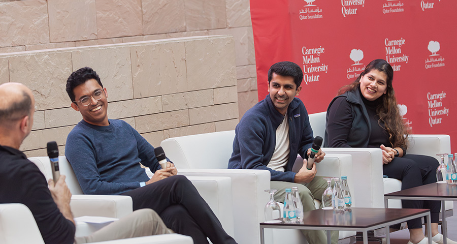  Two men and a woman sit in white chairs speaking to a man with a microphone.