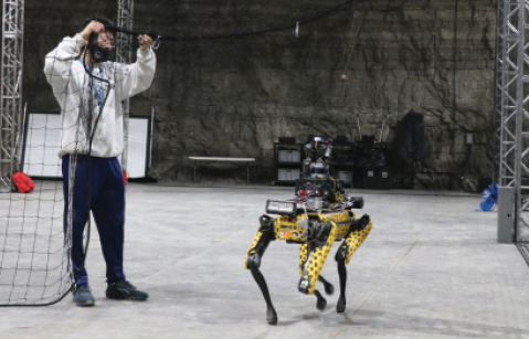 A man on the left of the photo interacts with a four-legged robot in an underground environment. The drone is yellow with black spots.