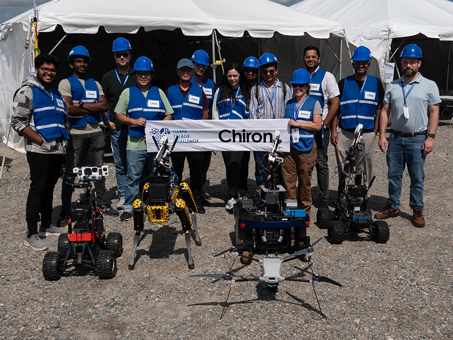  A team of men and women wearing blue vests and blue hardhats pose in front of a tent with four robots and a sign that says Chiron.