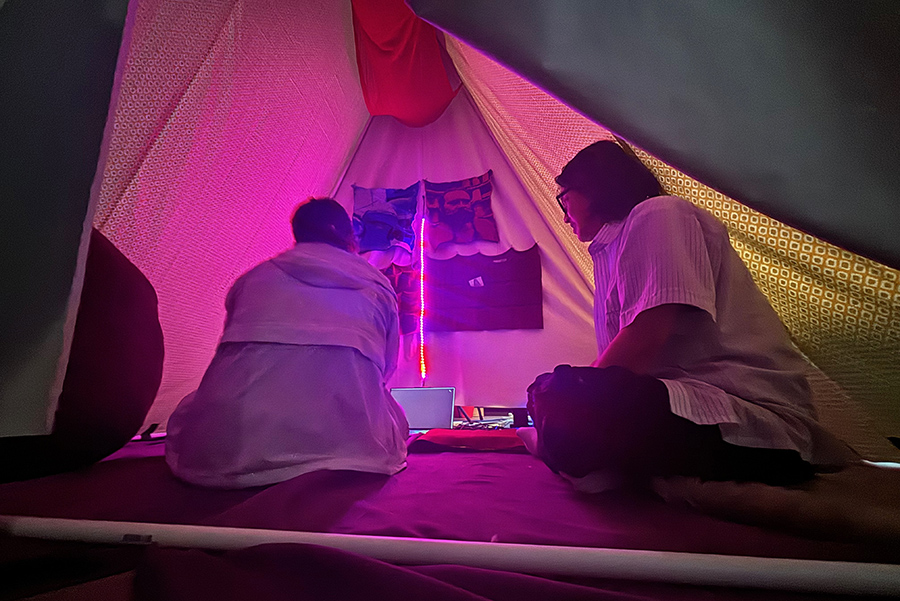  Two people sit inside a tent, whose lighting gives it a pink hue. Images of two people hang on one side of the tent, giving it a shrine-like feel.