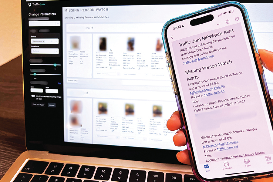  a hand holds a cell phone in front of a computer screen, both displaying the Missing Persons Watch software system from Marinus Analytics