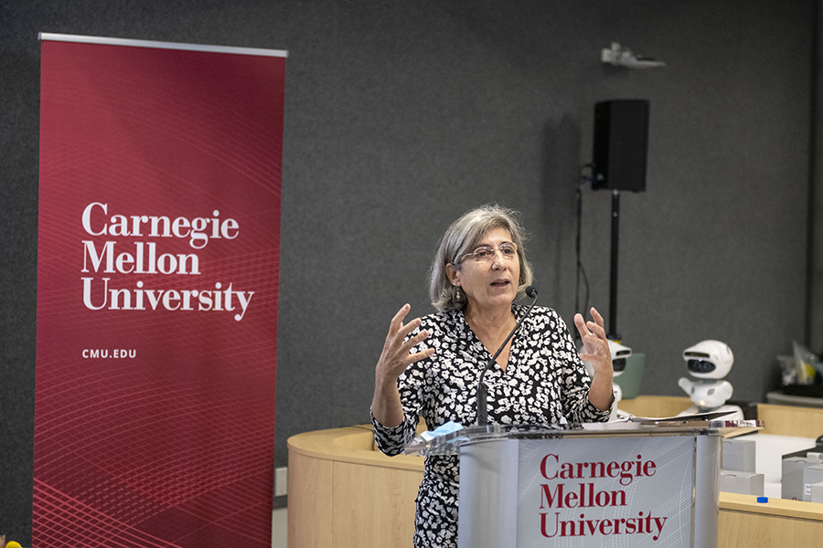 A woman with gray hair wearing a black and white dress stands behind a podium, speaking into a microphone. A Carnegie Mellon University banner is on the left, and a small white robot is in the background on the right.