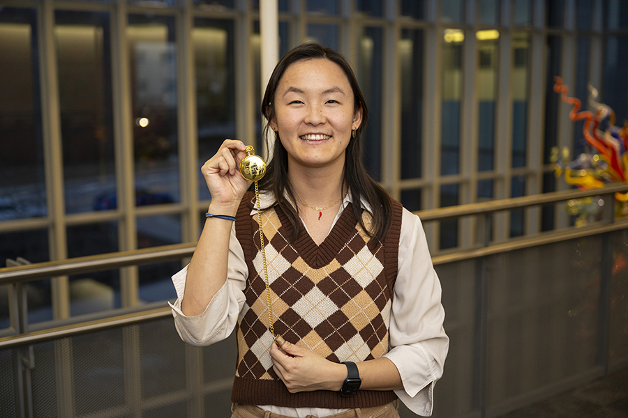  A woman in a brown and ivory argyle sweater smiles and holds a gold pocket watch up for the camera.