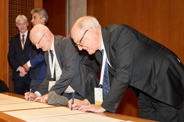  Two men lean over a table, signing a document.