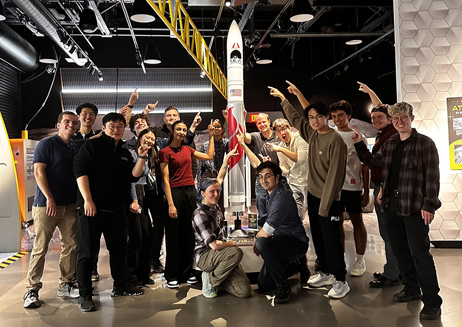  A group of students pose in front of a model of a lunar lander and point toward its nose, where a miniature rover they designed is stored.