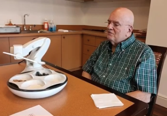  An older man in a plaid shirt sits at a table with a robotic arm that will help him eat.