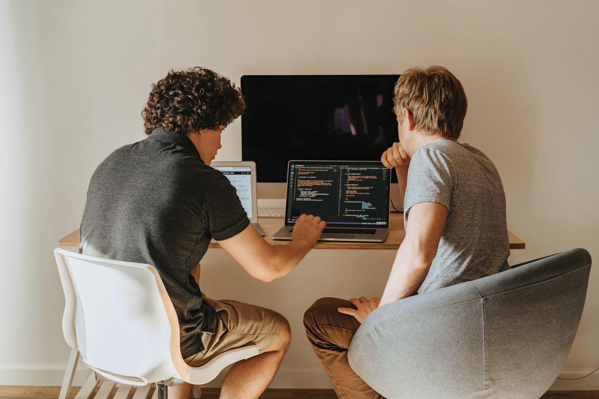 Two men sit in front of a laptop with their backs to the camera, engaged in what looks like computer programming.